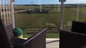 A balcony with wicker furniture and a view of a green field and distant buildings.