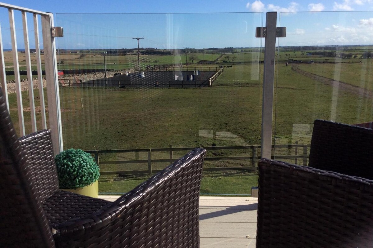 A balcony with wicker furniture and a view of a green field and distant buildings.