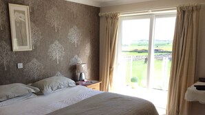 A bedroom with a large bed, a bedside table with a lamp, and a view of a green landscape through a sliding door.