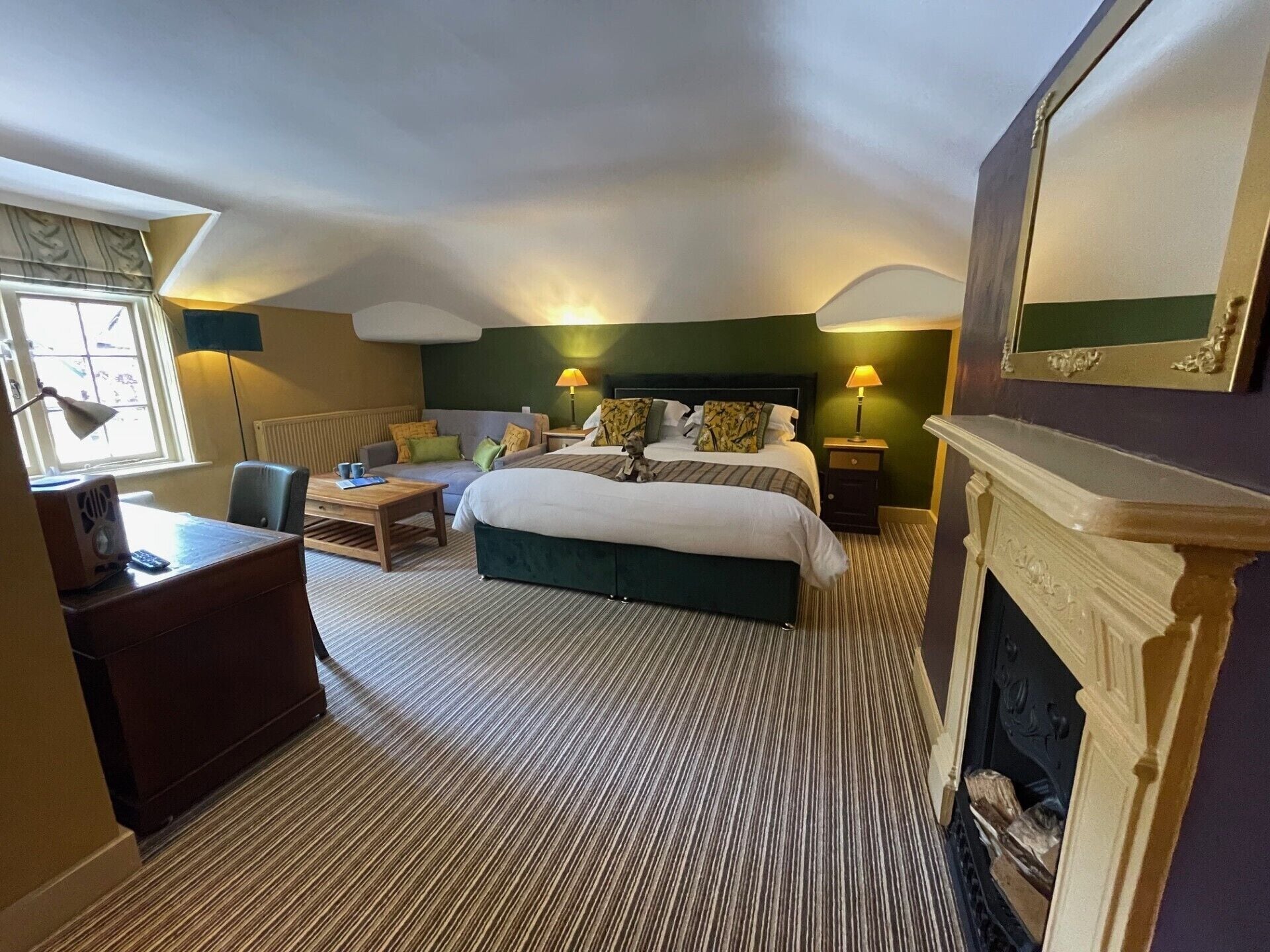 Una habitación de hotel con una cama grande, un escritorio, una silla, un sofá, dos mesitas de noche con lámparas, una chimenea y una ventana con cortinas.