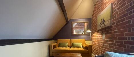 A cozy loft-style living room with a wooden floor, a yellow sofa with green cushions, a wooden coffee table, and a wicker basket.