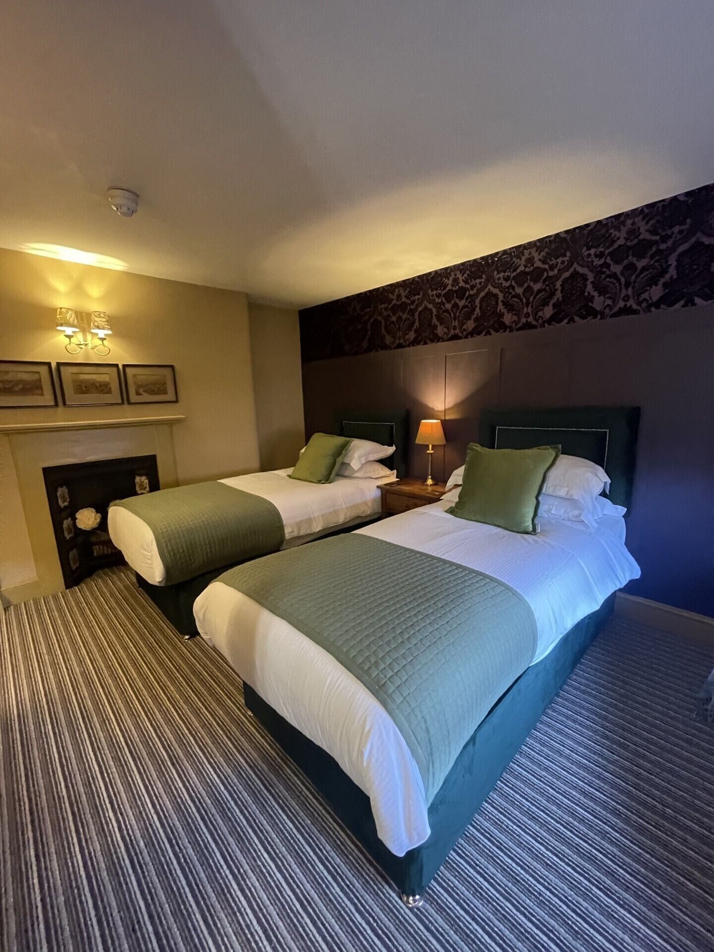 Une chambre d’hôtel avec deux lits, une cheminée, des lampes fixées au mur et un papier peint à motifs.