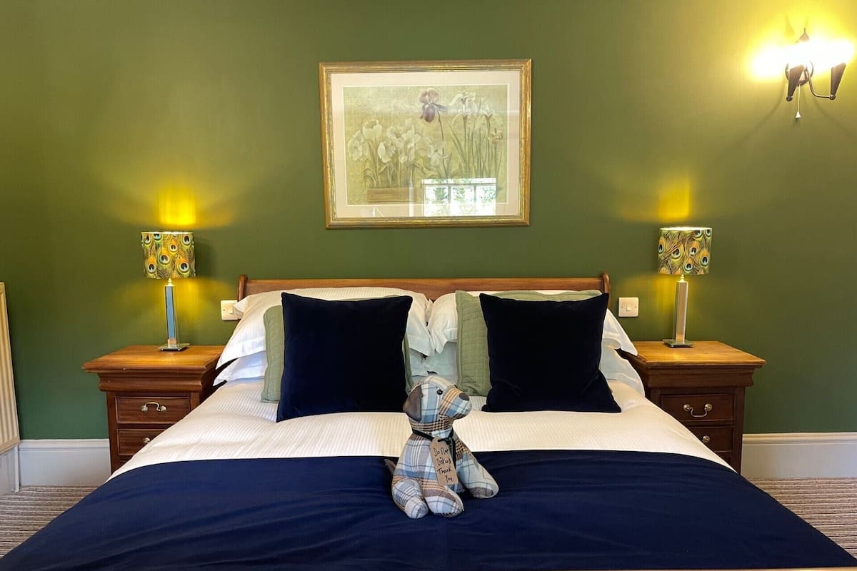 Cama bem arrumada com duas almofadas, um cobertor e um ursinho de pelúcia com uma mensagem. Há duas mesas de cabeceira com luminárias e uma moldura com um quadro na parede acima da cama.