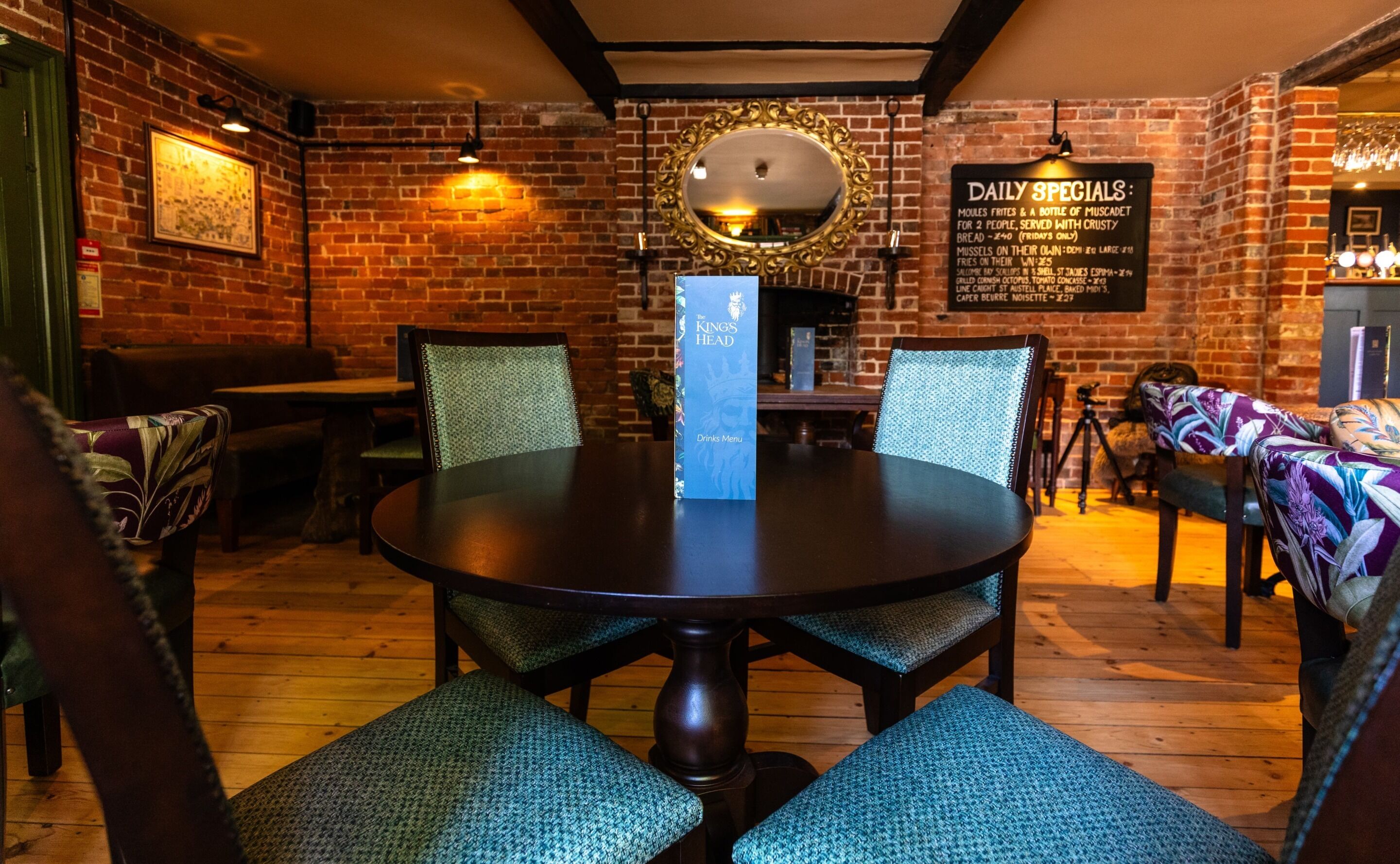 A restaurant with a brick wall, wooden tables, and chairs. There is a menu board on the wall and a mirror above the table.