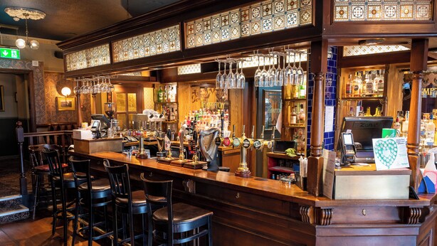 A bar area with wooden bar counters, multiple stools, and a variety of bottles and glasses. - The Hampton Hotel by Greene King Inns (Edinburgh)