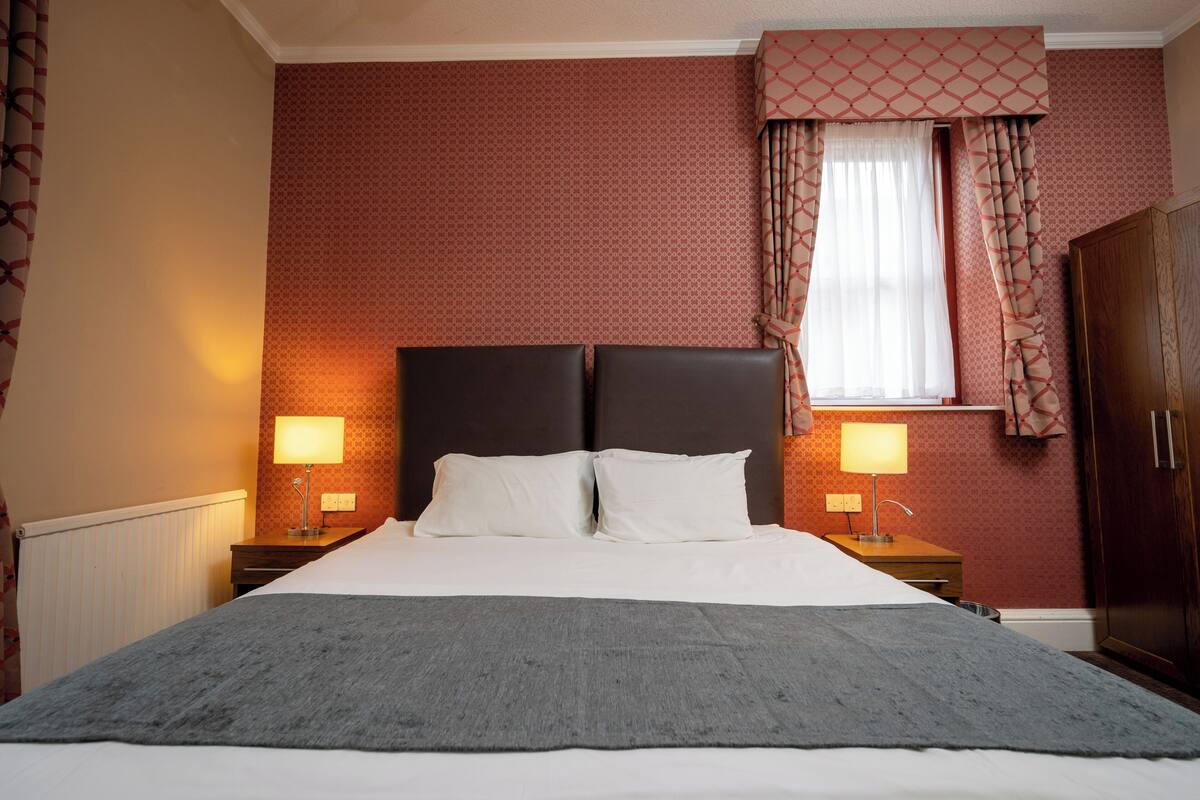 Una cama bien tendida con sábanas blancas y una manta gris, con dos lámparas de noche sobre mesitas de madera.