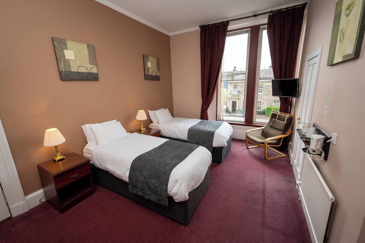 Una habitación de hotel con dos camas, una silla, un televisor y una ventana con cortinas.