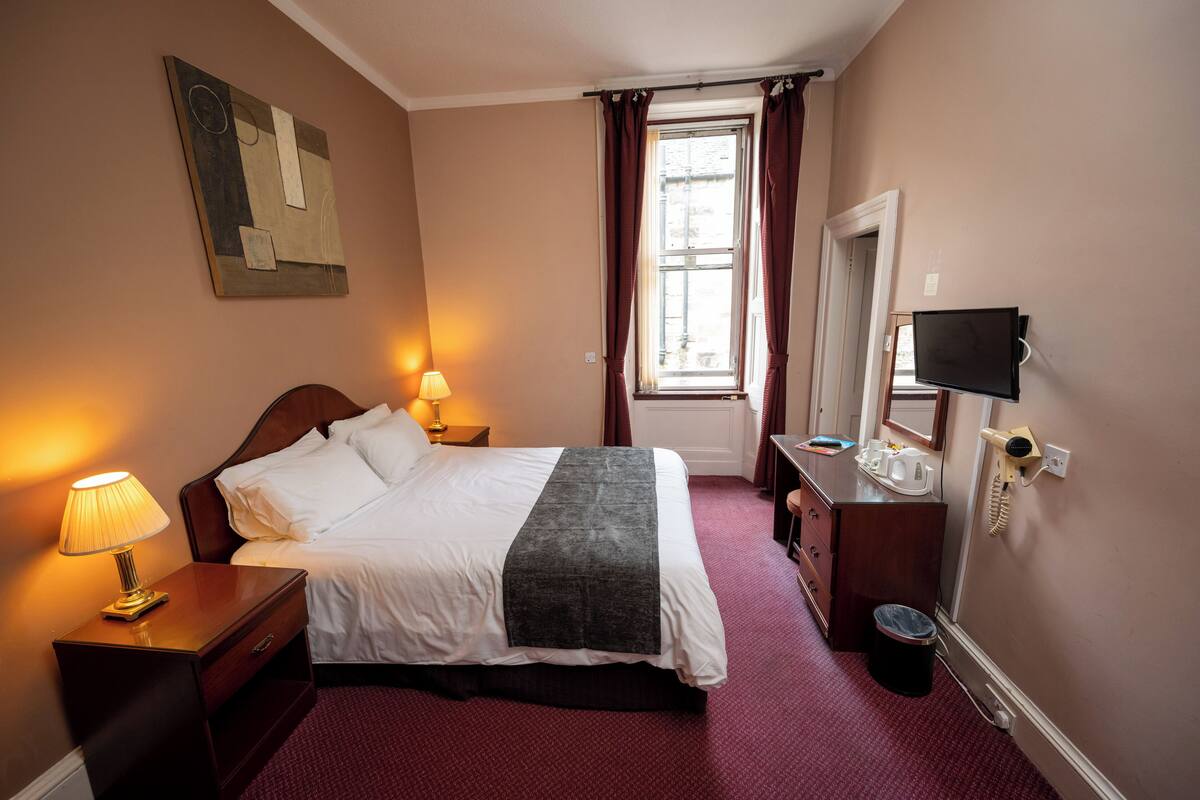 Habitación de hotel con cama, lámparas de noche, televisor, espejo y ventana con cortinas.