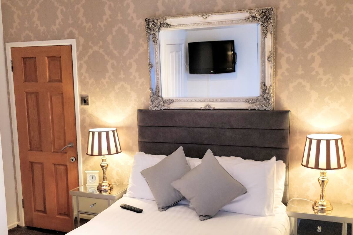 A hotel room with a large bed, two bedside tables with lamps, a television mounted above the bed, and a door to the left.