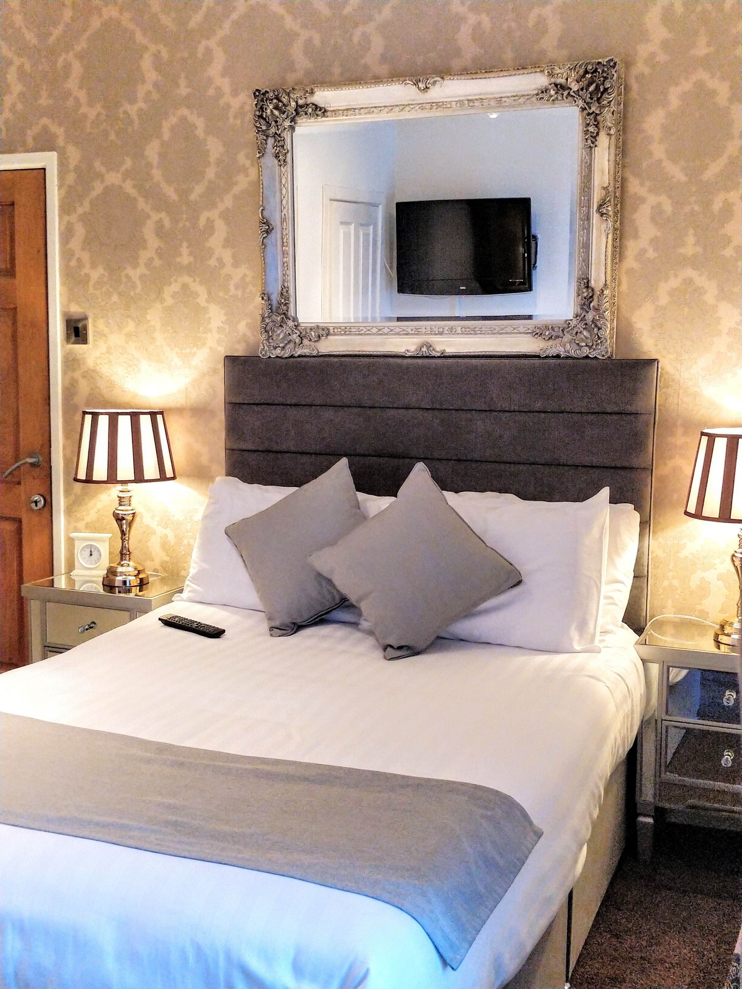 A neatly made bed with a headboard, two bedside lamps, a television above the bed, and a mirror with an ornate frame.