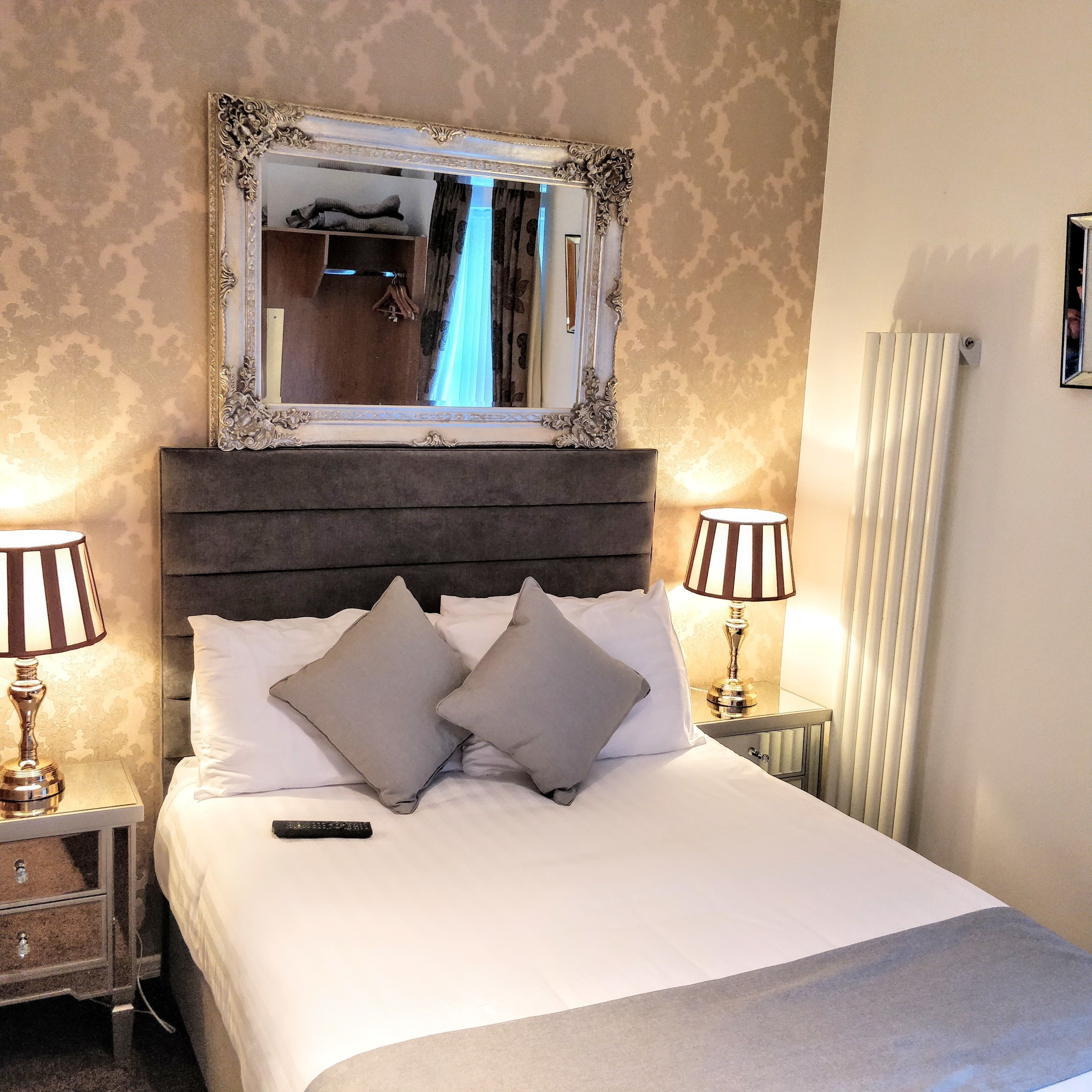 A neatly made bed with a large mirror above it, flanked by two bedside lamps, and a radiator on the wall.