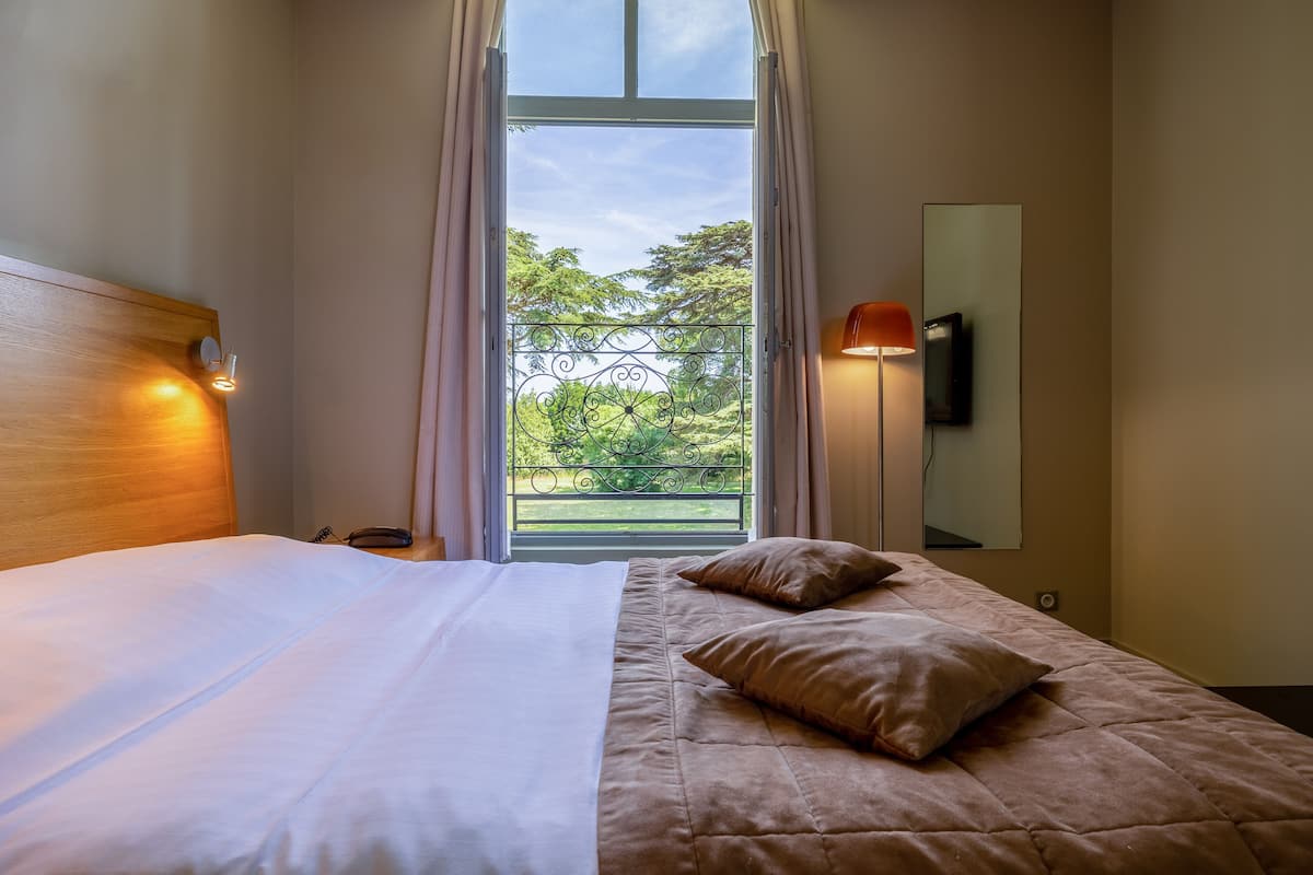 A hotel room with a bed, a nightstand, a telephone, a window with curtains, a balcony view, and a wall-mounted TV.