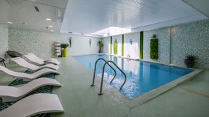 An indoor swimming pool with lounge chairs, a pool ladder, and tiled walls with green accents.