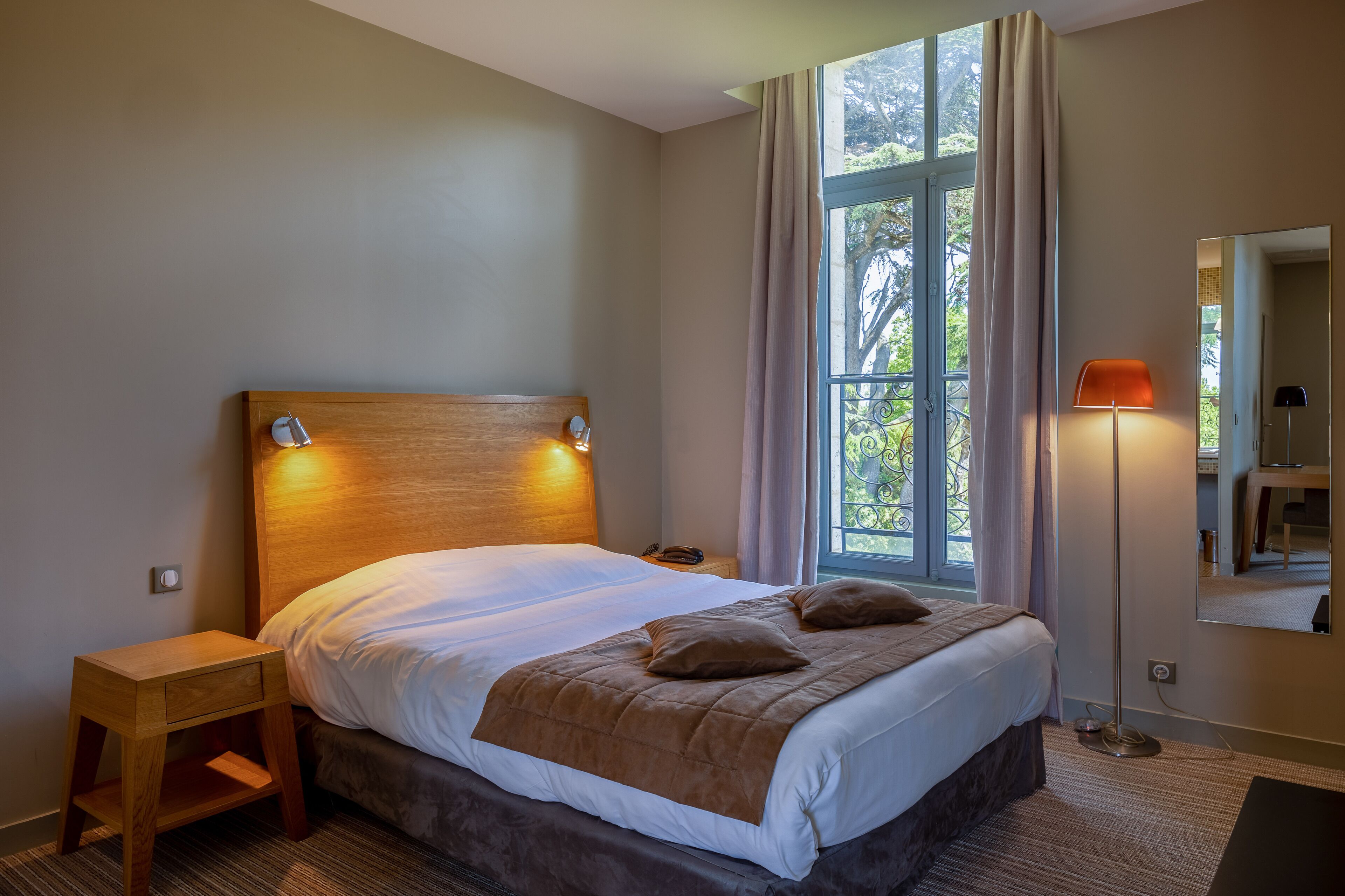 A neatly made bed with a wooden headboard, a bedside table with a drawer, a floor lamp, and a large window with curtains.