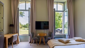 A hotel room with a bed, desk, chairs, a TV, and large windows with curtains.