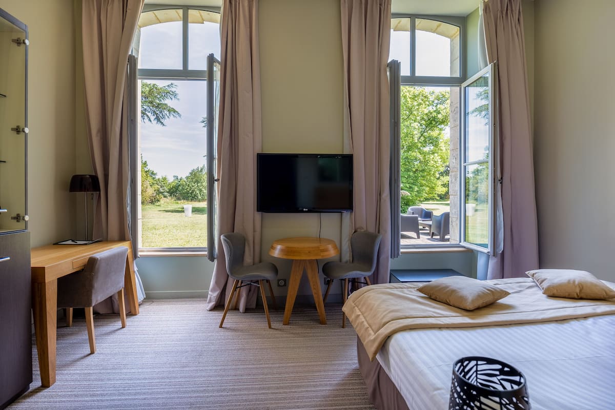A hotel room with a bed, desk, chairs, a TV, and large windows with curtains.
