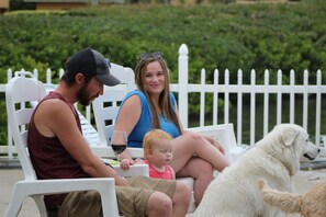 A family seated on white chairs outdoors with a white fence and greenery in the background.
