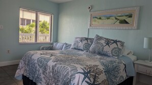 Une chambre à coucher avec un lit, une fenêtre avec des stores, un tableau accroché au mur et une table de chevet avec une lampe.