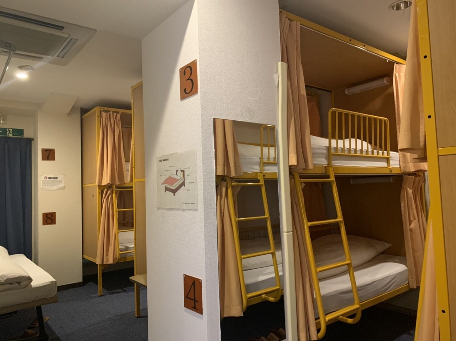 A dormitory room with bunk beds, numbered doors, a whiteboard with a diagram, and a small table with a mattress.
