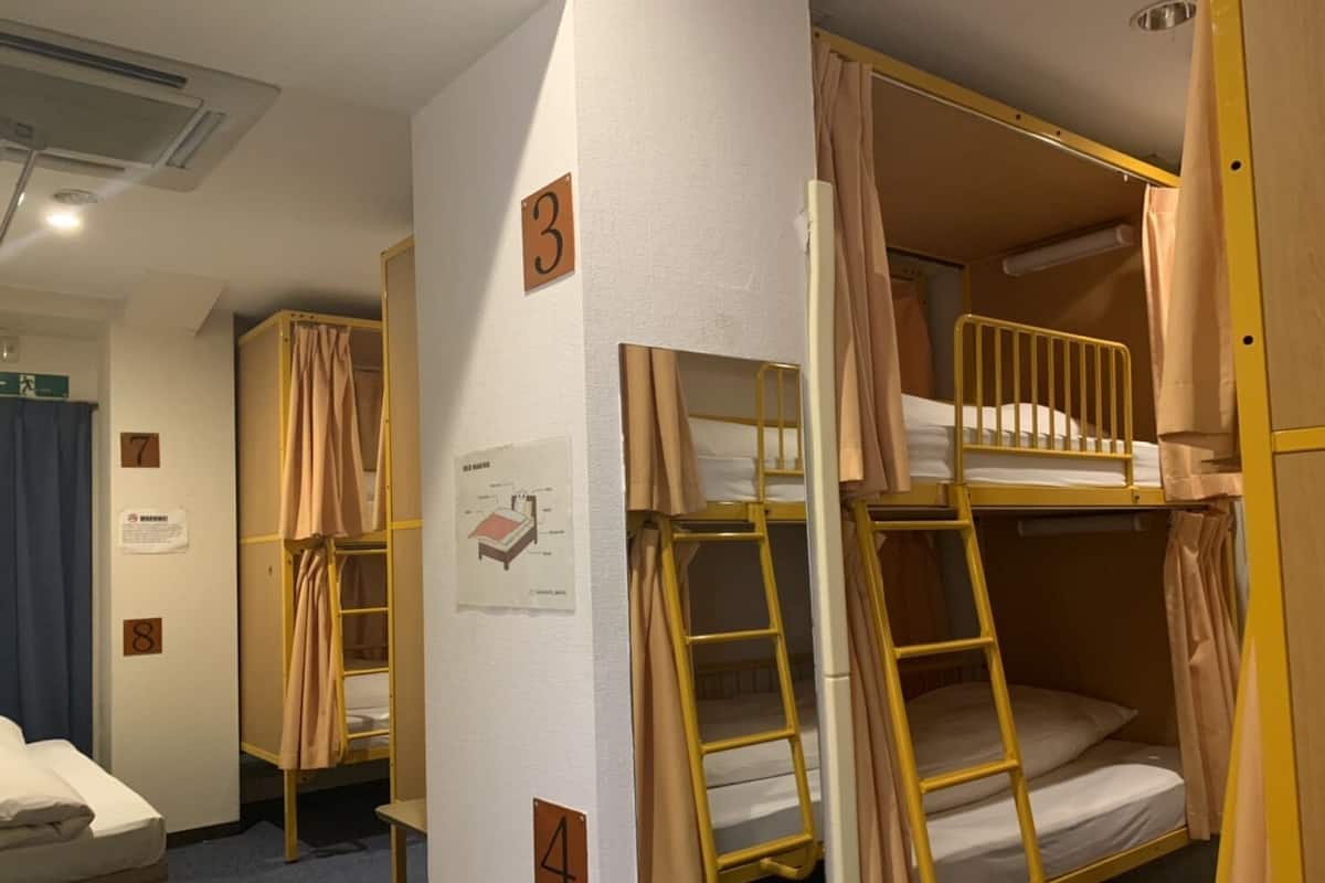 A dormitory room with bunk beds, numbered doors, a whiteboard with a diagram, and a small table with a mattress.