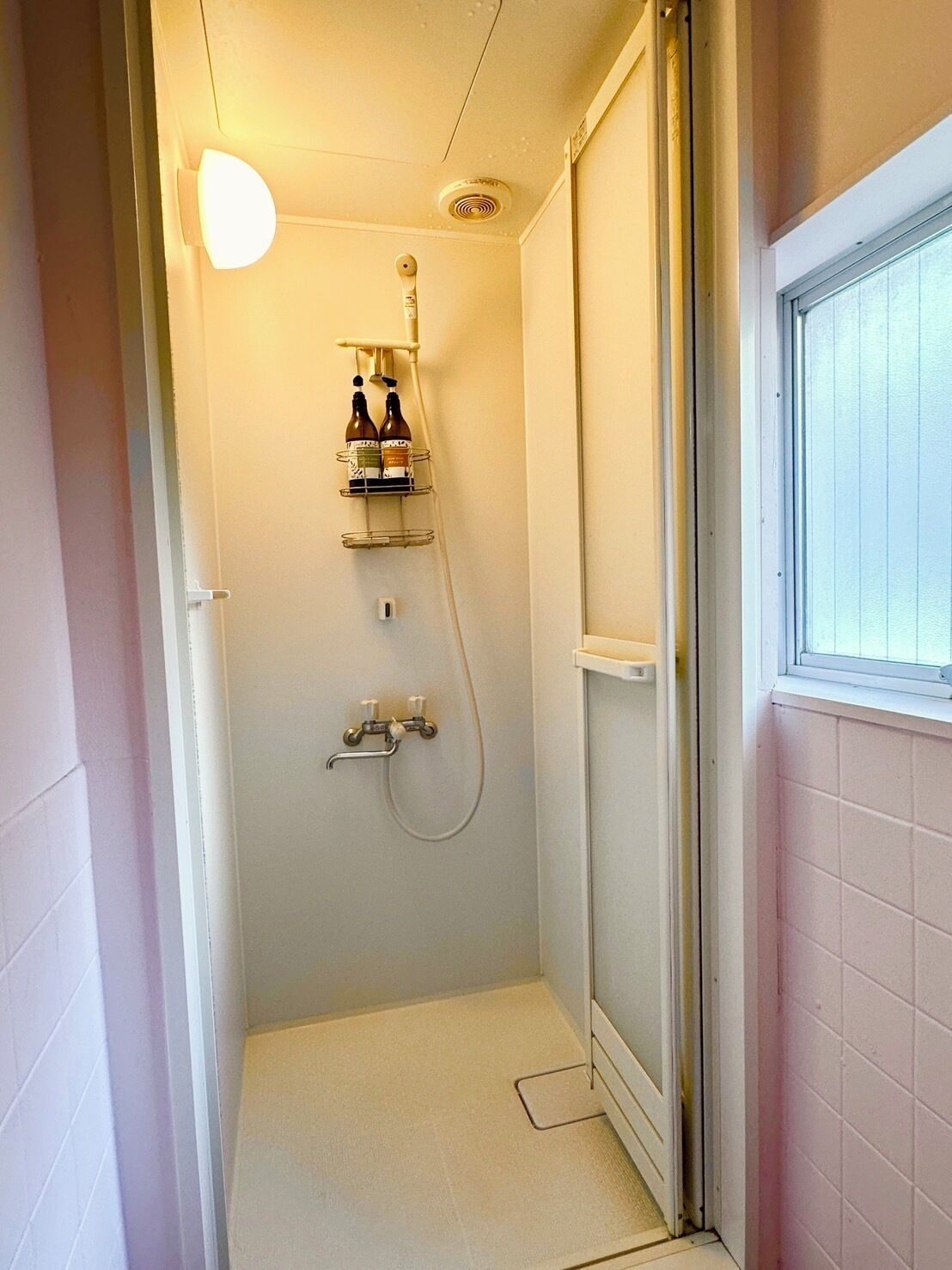 Een kleine, afgesloten douchecel met een glazen deur, een lamp boven de douche en een plank met flessen.