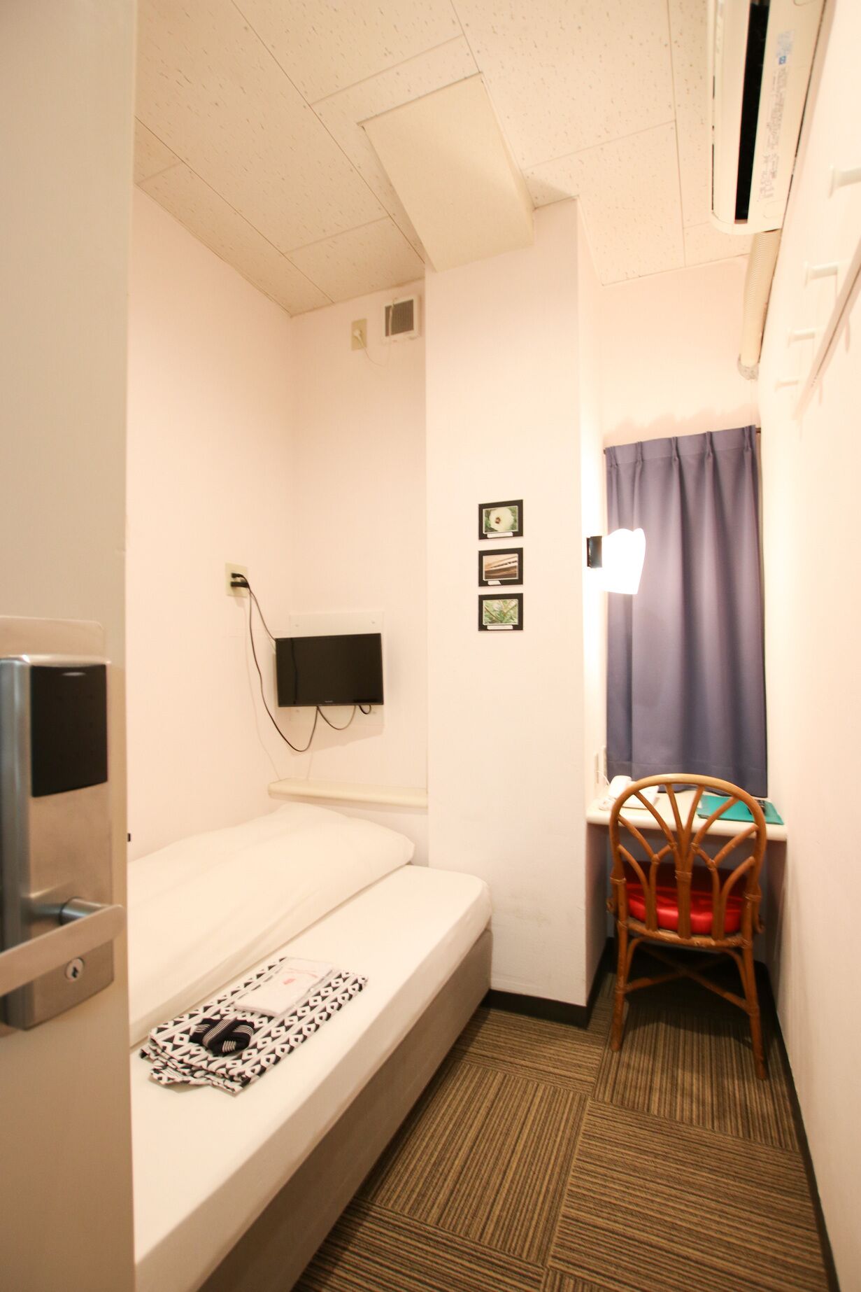 A small hotel room with a single bed, a chair, a TV mounted on the wall, and a window with curtains.
