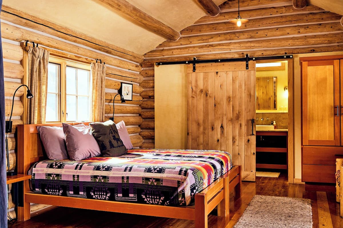 A log cabin bedroom with a wooden bed, a nightstand, a dresser, and a bathroom with a wooden vanity.