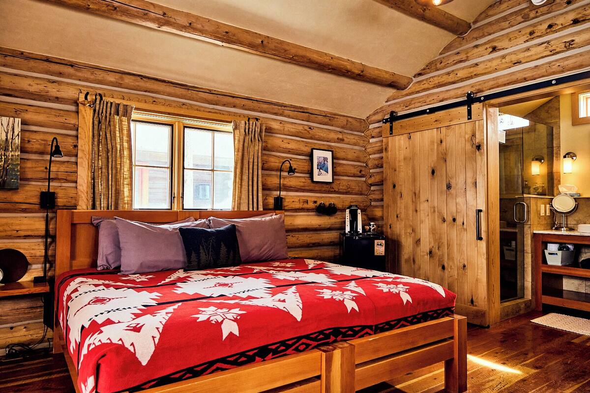 A wooden cabin-style bedroom with a bed, a nightstand, a dresser, and a large window with curtains.