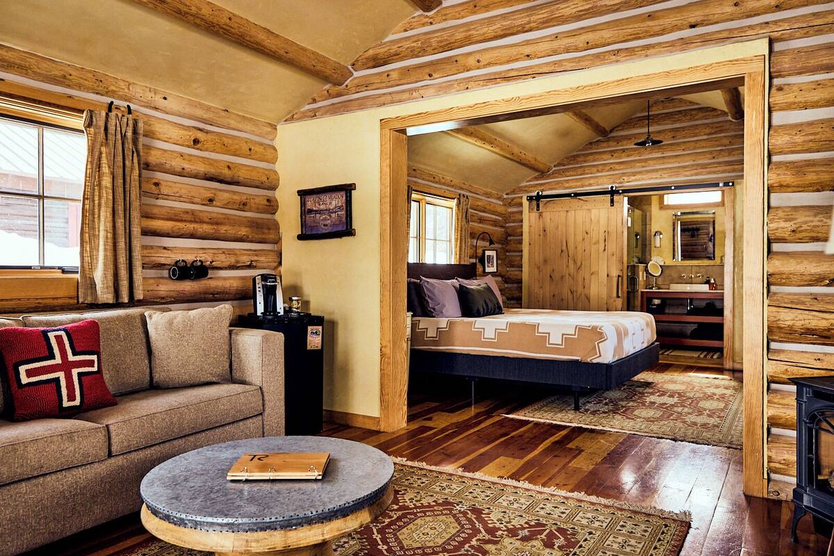 A log cabin interior with a sofa, coffee table, and a bed with a patterned bedspread.