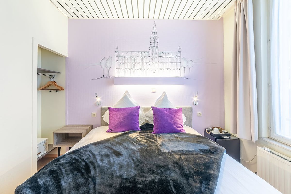 A hotel room with a bed, two purple pillows, a headboard with a light fixture, and a window with curtains.