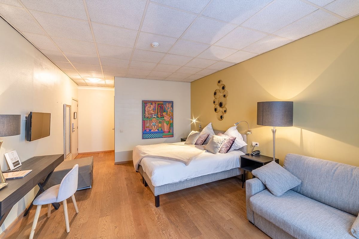 A modern hotel room with two beds, a desk, a sofa, and a TV.