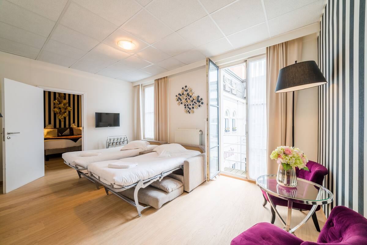 A modern hotel room with a bed, a small table with a vase, a purple chair, and a view of the city through the window.