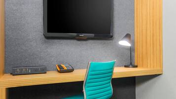 A modern room with a flat-screen TV mounted on a grey textured wall, a wooden desk with a teal chair, and a desk lamp.