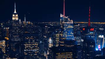 Une vue nocturne d’une ville, avec des gratte-ciel illuminés, dont l’Empire State Building et un immeuble surmonté d’une flèche rouge.
