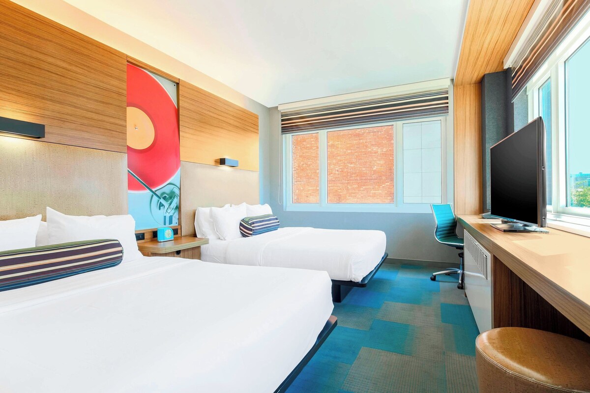 Een hotelkamer met twee bedden, een bureau met een televisie, een stoel en een groot raam met uitzicht op de stad.