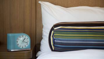 A bed with white bedding and a striped pillow, a blue clock on a wooden nightstand, and a wooden headboard.