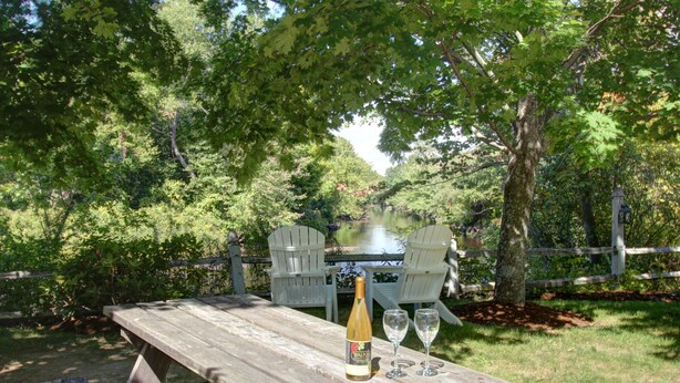 A wooden picnic table with a bottle and two glasses, surrounded by lush greenery and a wooden fence. - Riverhouse by Weekender (Formerly Jack Daniels Inn) (Peterborough)