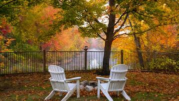 Two white Adirondack chairs facing each other with a tree in the background and a fence enclosing the area.