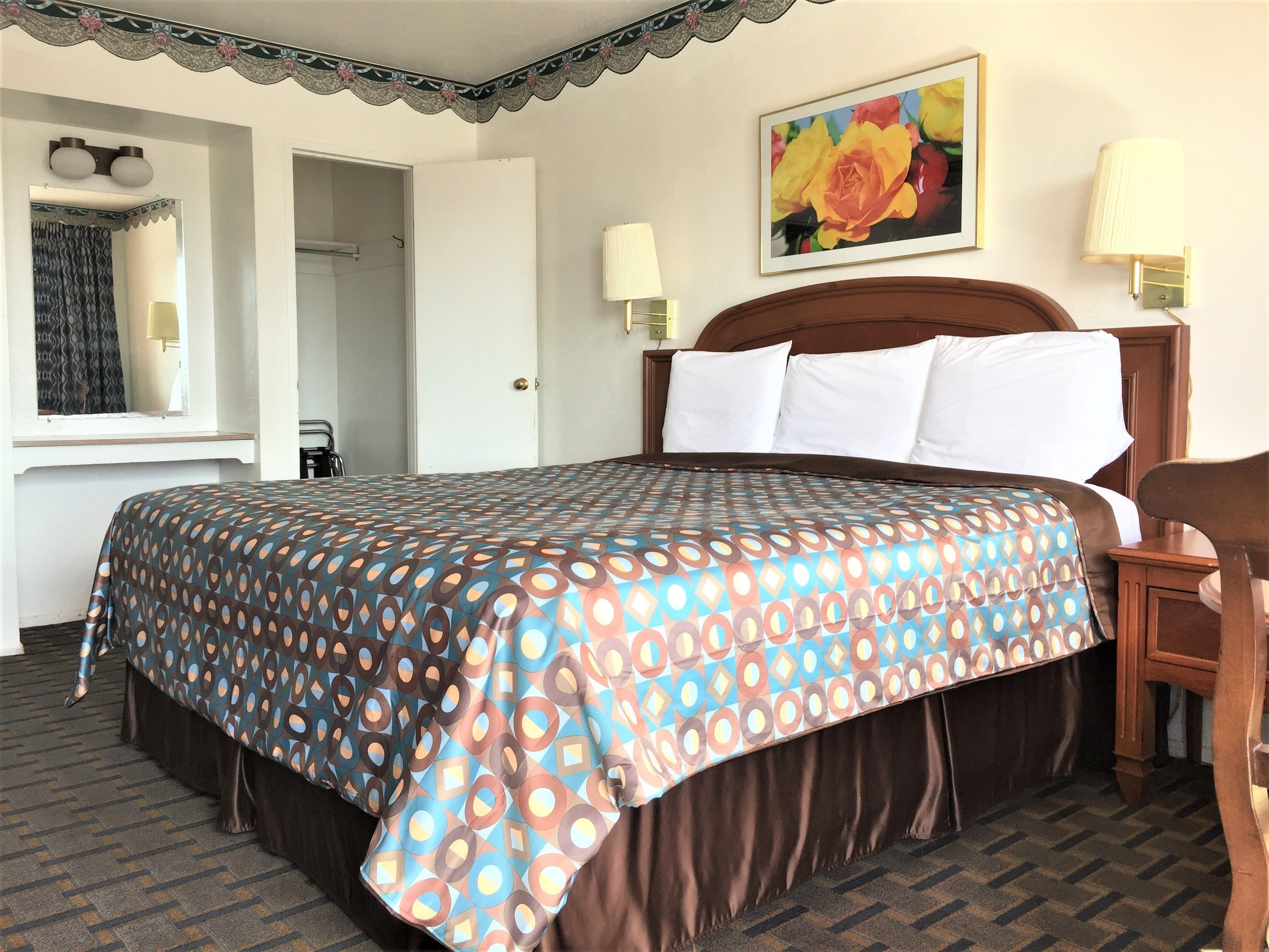 A hotel room with a large bed, two bedside lamps, a painting of flowers, a mirror, and a window with curtains.