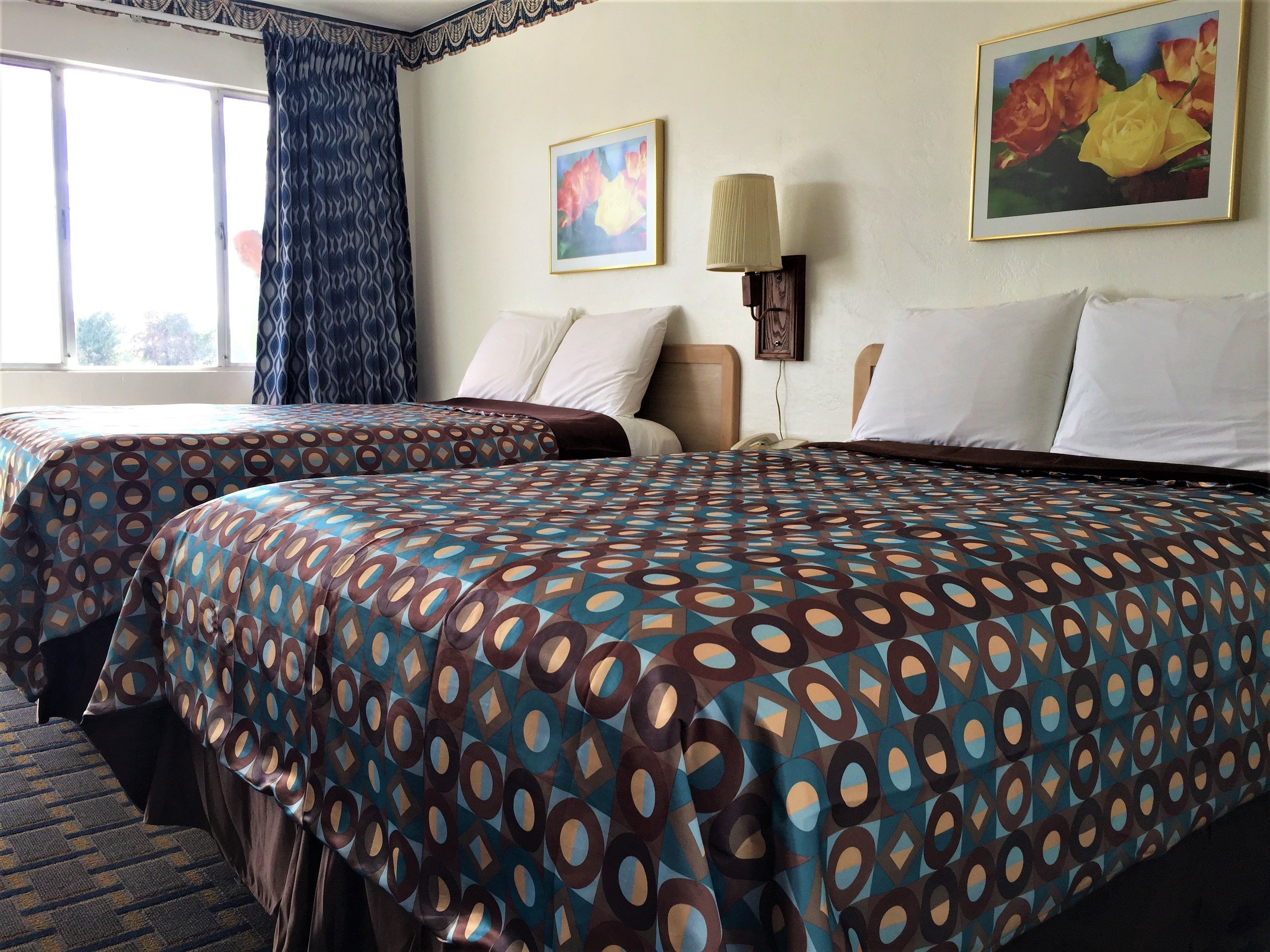 Une chambre d’hôtel avec deux lits, des couvre-lits à motifs, une fenêtre avec des rideaux et deux tableaux au mur.