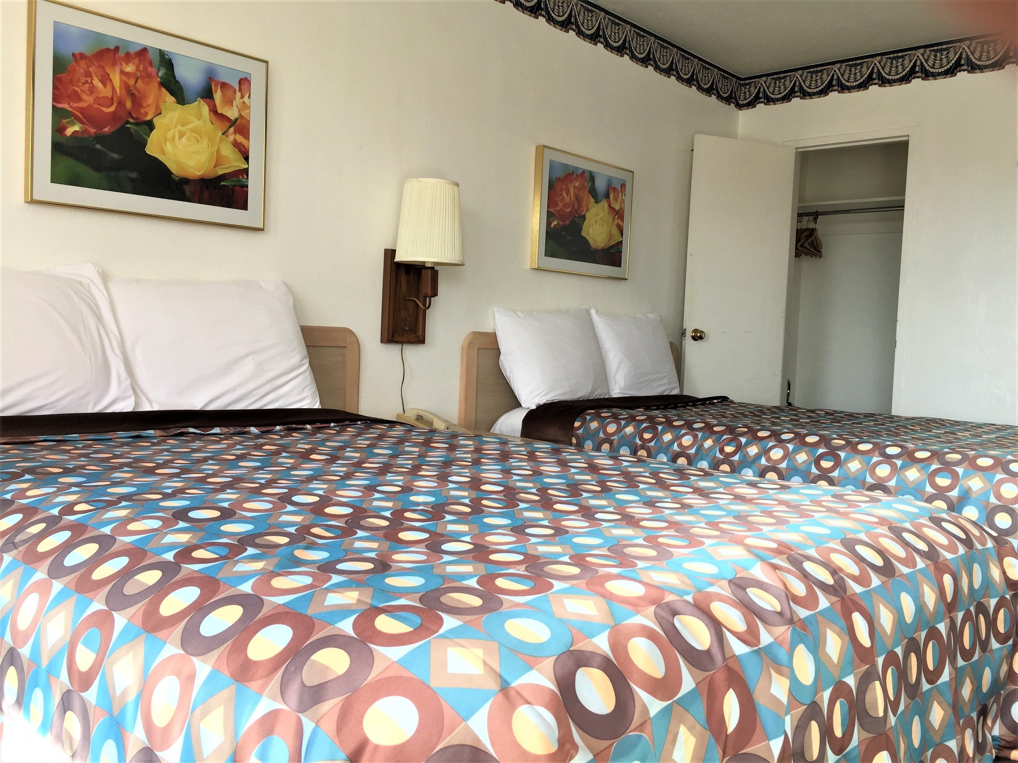 Quarto de hotel com duas camas, cada uma com colcha estampada, almofadas brancas e mesa de cabeceira com abajur. Há duas quadros nas paredes, e uma porta que dá para um armário.