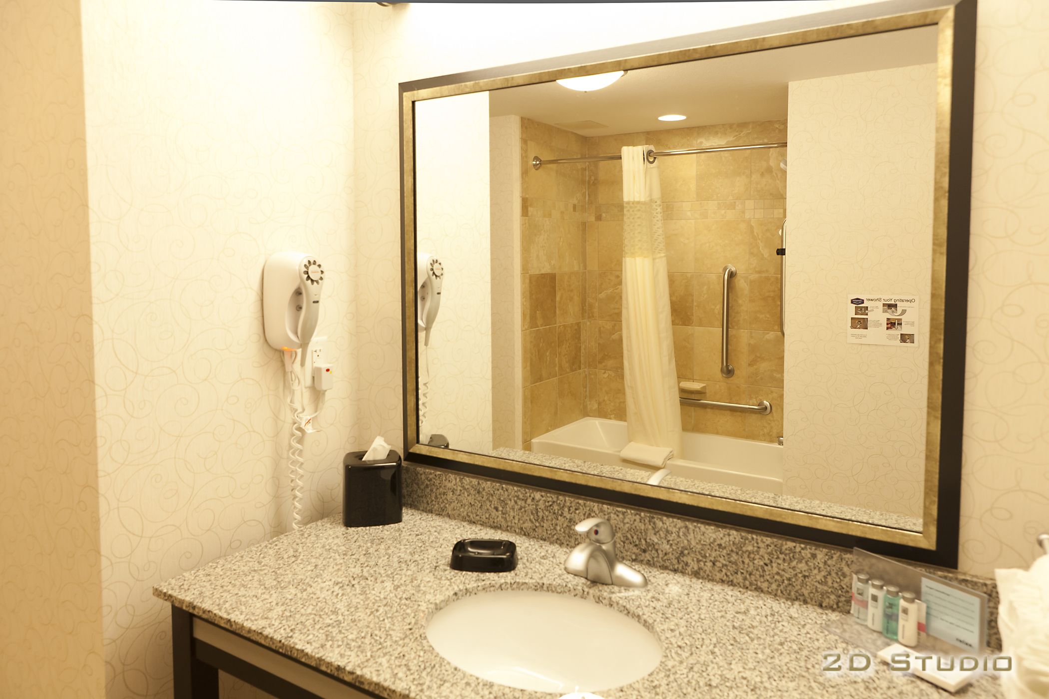 Bany amb taulell de granit, un lavabo, un mirall, un dispensador de tovalloles i una dutxa visible al reflex.