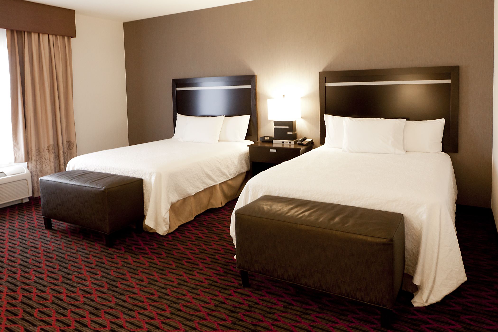 Une chambre d’hôtel avec deux lits, une table de chevet avec une lampe, une fenêtre avec des rideaux et un tapis à motifs.