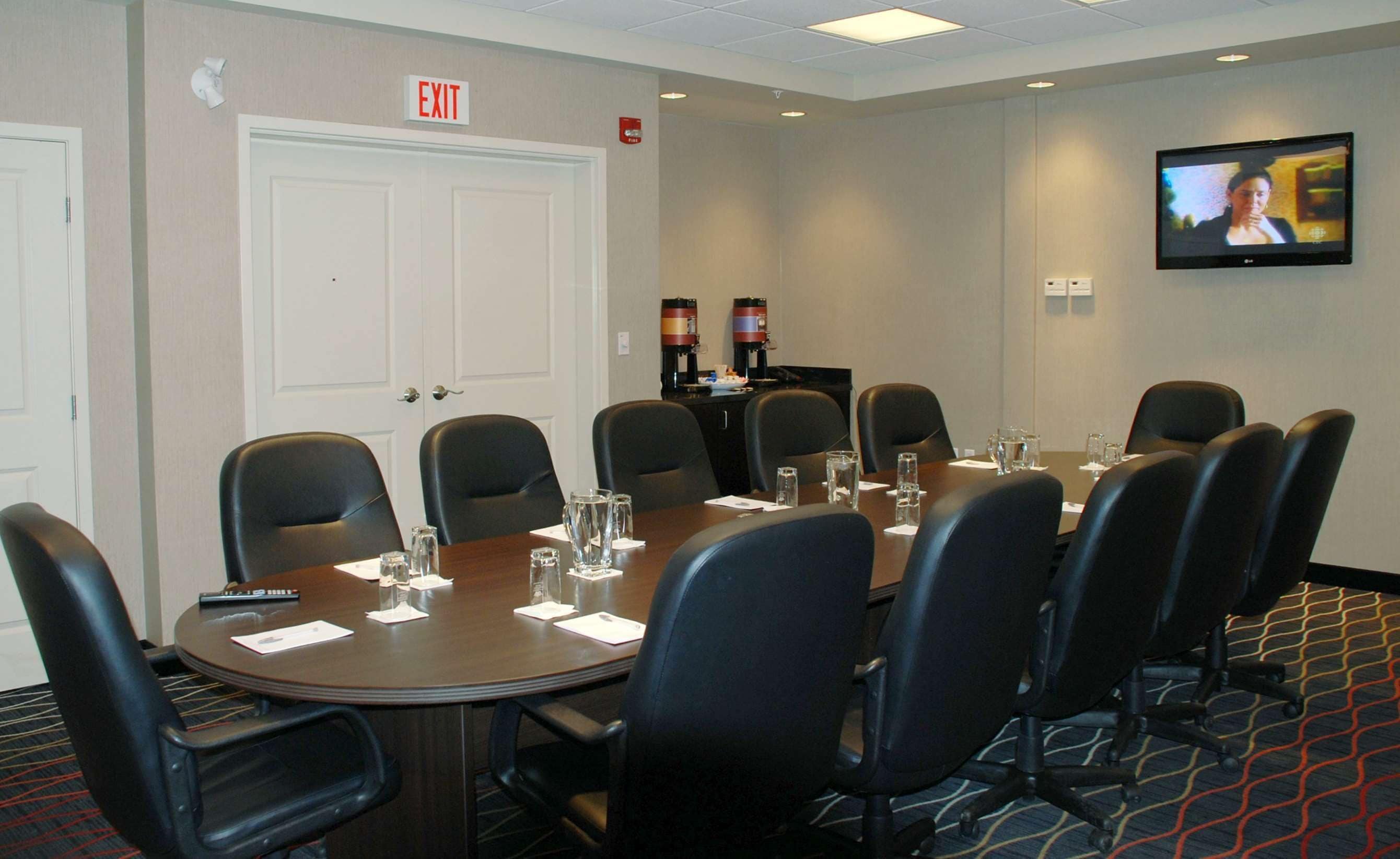 A conference room with a large table, black chairs, a TV on the wall, and an exit sign.