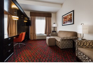 Un cuarto de hotel con cama, escritorio, sillón, sofá, lámpara, un cuadro en la pared y ventana con cortinas.