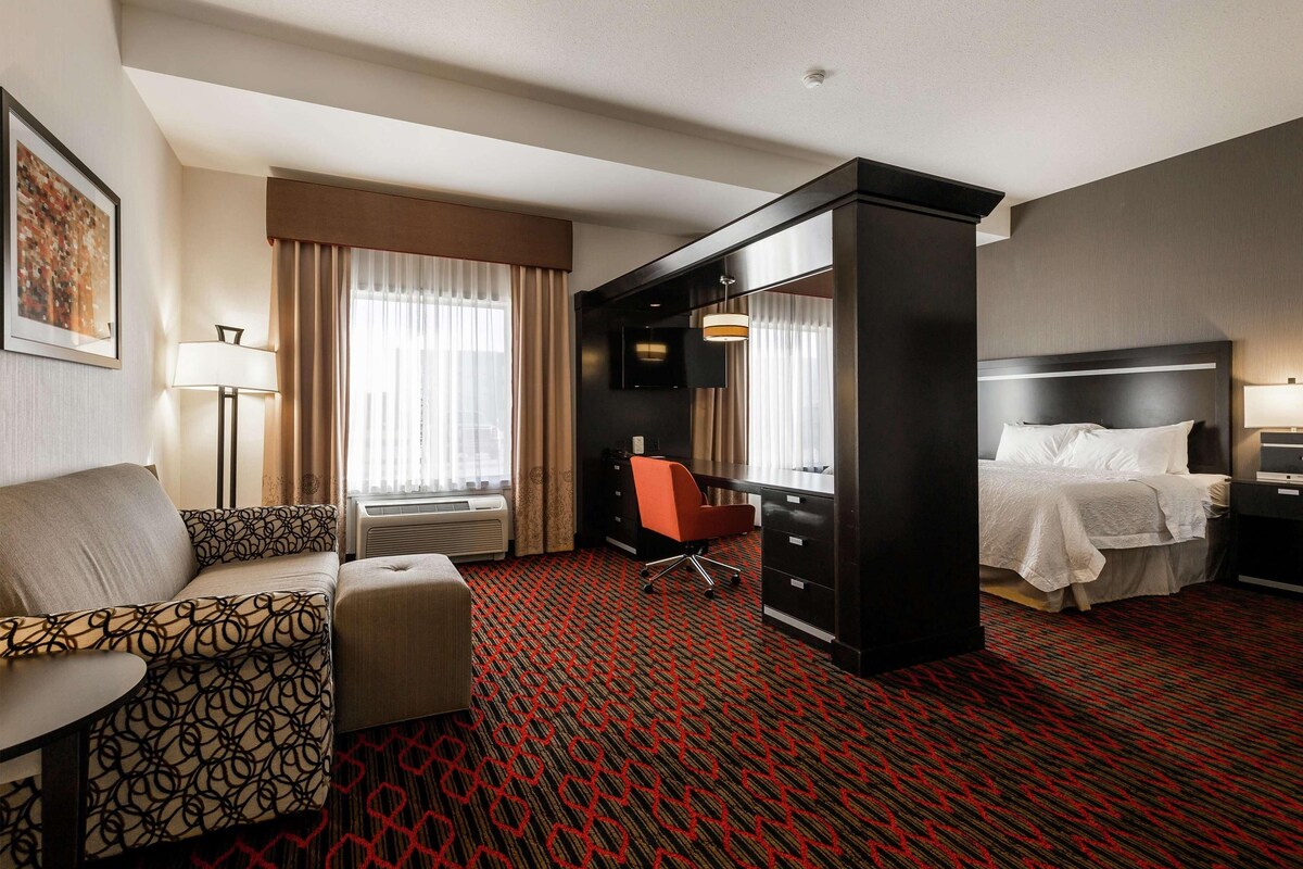 Una habitación de hotel con una cama, un escritorio, una silla, un sofá y un ventanal con cortinas.