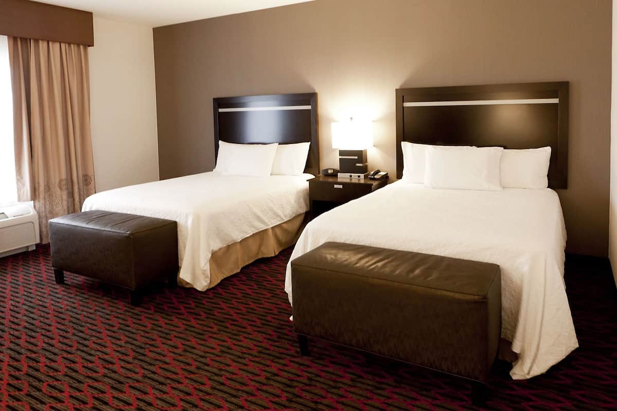 A hotel room with two beds, a nightstand, a lamp, and a window with curtains.