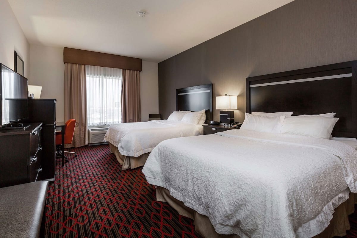 A hotel room with two beds, a desk, a chair, a TV, and a window with curtains.