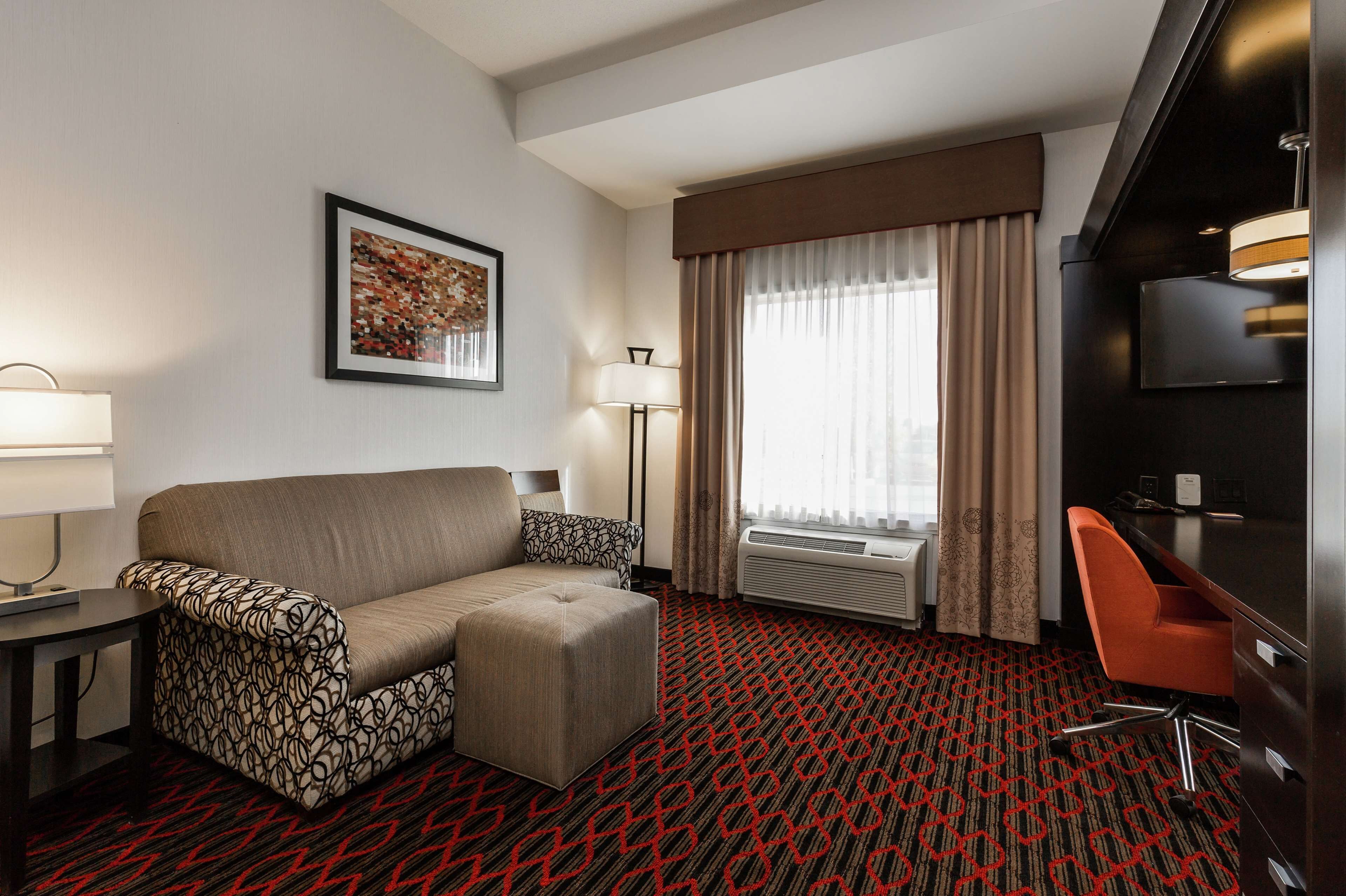 A hotel room with a sofa, ottoman, desk, chair, air conditioning unit, and a television.