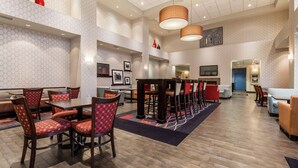 Un amplio vestíbulo de hotel con techos altos, varias zonas de estar, incluyendo una larga mesa de comedor y cabinas, y un televisor montado en la pared.